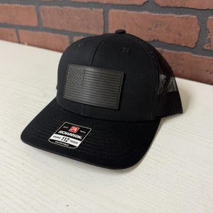 Puede incluir: Gorra de camionero negra con parte trasera de malla y visera curva. La gorra presenta un parche rectangular con un diseño de bandera americana. La etiqueta de la marca "Richardson 112 Trucker" es visible.