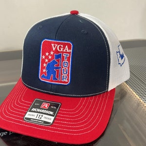 May include: A blue and red trucker hat with a white mesh back. The hat has a red, white, and blue patch with the text "VGA TOUR" and a cartoon soldier holding a flag. The hat is made by Richardson and is adjustable.