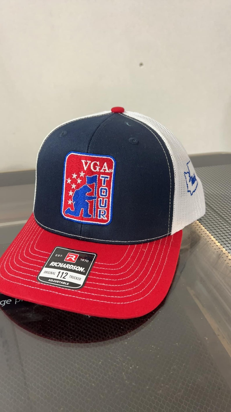 May include: A blue and red trucker hat with a white mesh back. The hat has a red, white, and blue patch with the text "VGA TOUR" and a cartoon soldier holding a flag. The hat is made by Richardson and is adjustable.