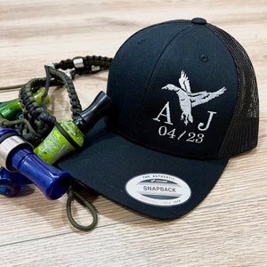 May include: Black and white embroidered trucker hat with a flying duck and the initials "AJ" with the date "04/23". The hat has a black mesh back and a black front with a white "The Authentic Snapback" label.