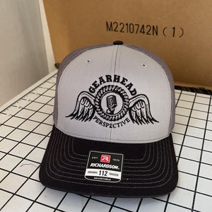 May include: Gray and black trucker hat with a black and white logo that says "Gearhead Perspective" with a microphone and wings. The hat is made by Richardson 112.