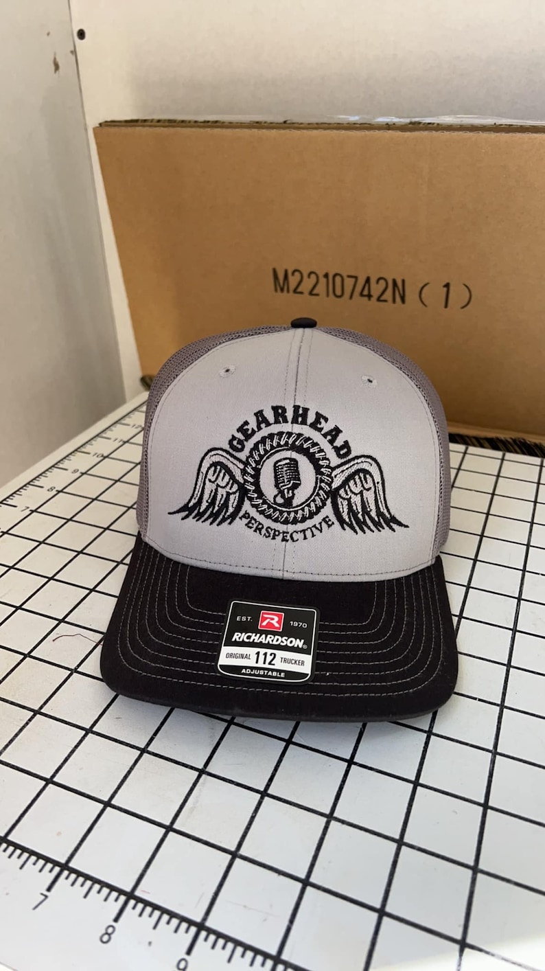 May include: Gray and black trucker hat with a black and white logo that says "Gearhead Perspective" with a microphone and wings. The hat is made by Richardson 112.