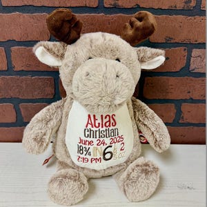 May include: A plush moose toy in shades of brown and tan, with a white bib embroidered with the name "Atlas", birthdate, and measurements. The moose has brown antlers and a soft, furry texture, perfect for a baby.