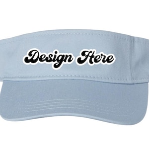 Puede incluir: Visera azul claro con el texto "Design Here" en negro, con una fuente cursiva. La visera tiene una visera curvada y una banda. Este accesorio está diseñado para proteger los ojos del sol.