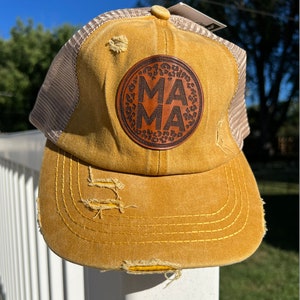 May include: A yellow distressed trucker hat with a brown leather patch that says "MAMA" in a circle with a leopard print design.
