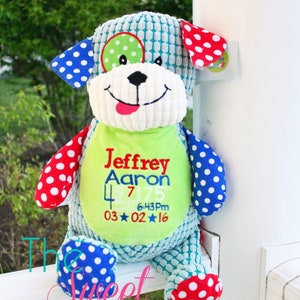May include: A plush blue, green, red, and white dog toy with a personalized embroidered name, date, and time. The toy is sitting on a white railing.