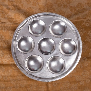 May include: A silver metal baking pan with seven round depressions. The pan is on a brown fabric background.
