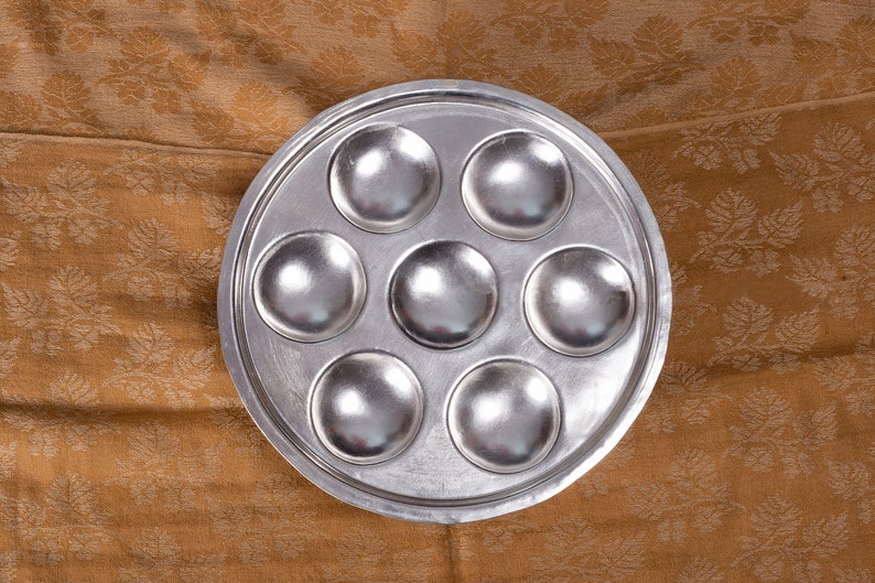 May include: A silver metal baking pan with seven round depressions. The pan is on a brown fabric background.