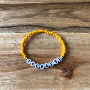 Peut inclure: Un bracelet en perles jaunes avec des perles de lettres blanches qui épellent "J'aime le fromage" avec une perle de cœur rouge.