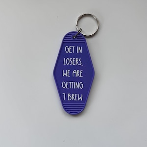 May include: A purple motel-style keychain with the phrase "GET IN LOSERS, WE ARE GETTING 7 BREW" in white lettering. The keychain has a silver metal ring attached at the top.