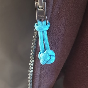 May include: Close-up of a silver zipper with a bright blue cord pull. The zipper is attached to a dark purple fabric. The zipper pull is knotted at the end.