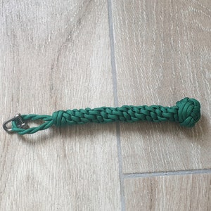 May include: A green macrame keyring with a metal ring. The keyring is made of woven green cord, with a knot at one end. The keyring is approximately 15 cm long.