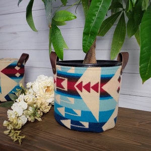 Könnte beinhalten: Zwei Stoff-Pflanztöpfe mit Ledergriffen. Die Töpfe haben ein geometrisches Muster in Blautönen, Rot und Weiß. Der größere Topf ist mit einer grünen Pflanze gefüllt.