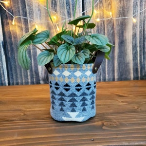 Könnte beinhalten: Eine kleine Topfpflanze in einem blauen und weißen Stofftopf mit Ledergriffen. Der Topf hat ein geometrisches Muster mit Dreiecken und Rauten.