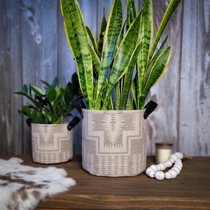 Könnte beinhalten: Zwei Topfpflanzen in Stoffgefäßen mit geometrischen Mustern. Der größere Topf ist beige mit einem braunen geometrischen Muster und der kleinere Topf ist beige mit einem grauen geometrischen Muster. Beide Töpfe haben schwarze Ledergriffe.