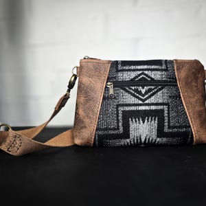 Puede incluir: Un bolso bandolera de cuero marrón con una solapa de tela estampada en blanco y negro. El bolso tiene cierre de cremallera y una correa larga ajustable.