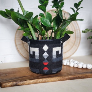 Könnte beinhalten: Ein schwarzer und weißer gemusterter Pflanzer mit Ledergriffen, gefüllt mit einer grünen Pflanze. Der Pflanzer steht auf einem Holzbrett.