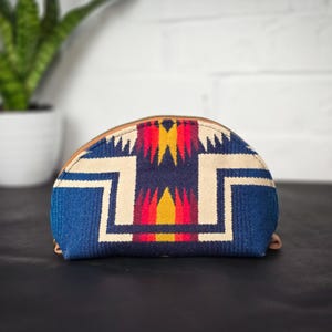 May include: A half-moon shaped blue and white patterned cosmetic bag with red and yellow accents. The bag has a zipper closure and a brown leather trim.