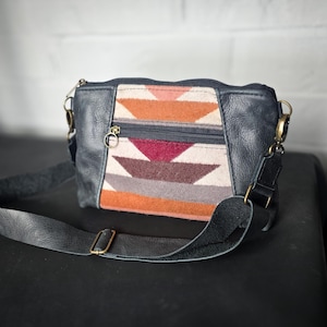 Puede incluir: Un bolso bandolera de cuero negro con un panel frontal con estampado geométrico colorido. El bolso tiene un compartimento principal con cremallera y una correa larga y ajustable. Los herrajes dorados acentúan el diseño del bolso.