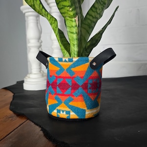 Könnte beinhalten: Ein farbenfroher, gemusterter Pflanztopf mit schwarzen Ledergriffen. Der Topf ist mit einer grünen Schlangenpflanze gefüllt.