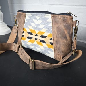 Puede incluir: Un bolso de cuero marrón cruzado con una solapa de tela con estampado geométrico. El bolso tiene una correa larga ajustable y un cierre de cremallera.