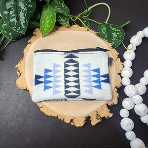 May include: A small white zippered pouch with a blue and black geometric pattern. The pouch is sitting on a wooden slice.