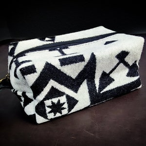 May include: A black and white geometric patterned toiletry bag with a zipper closure and a black leather strap.
