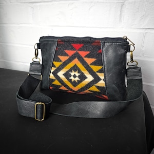 Puede incluir: Un bolso bandolera de cuero negro con una solapa de patrón geométrico. La solapa está hecha de una tela tejida colorida con formas geométricas rojas, naranjas, amarillas y negras.