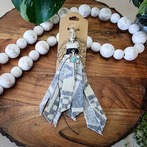 May include: A gray and white patterned tassel keychain with a silver charm featuring a turquoise stone. The keychain is hanging from a wooden bead garland.