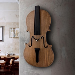 May include: Wooden violin-shaped wall clock with a natural wood finish. The clock features black details for the fingerboard, bridge, and scroll. The clock hands are black, and the clock is mounted on a gray wall.