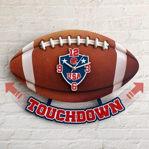 May include: A football-shaped wall clock with a brown and white design. The clock face features a blue shield with the text "USA" and a football helmet. The word "TOUCHDOWN" is displayed below the clock.