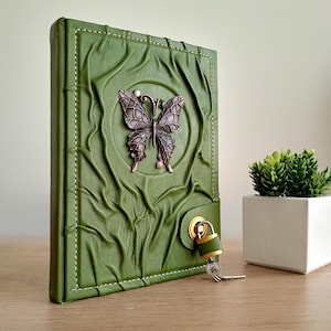 May include: Green leather journal with a butterfly embellishment. The journal has a textured cover with a raised butterfly design in the center. A gold-colored lock and key secure the journal. A small plant in a white pot is in the background.