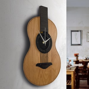 Peut inclure: Une horloge murale en bois en forme de guitare acoustique. Le cadran noir est orné d'aiguilles argentées, tandis que le corps de la guitare est en bois brun clair. L'horloge est fixée sur un mur gris clair.
