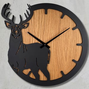 May include: A round wall clock featuring a natural light brown wood grain face and a black metal frame. A black silhouette of a deer with large antlers is integrated into the left side of the clock, with black hour and minute hands.