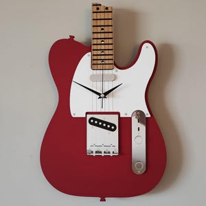 May include: A red and white guitar-shaped wall clock. The clock features a wooden neck and fretboard, with black clock hands. The body of the guitar is red, with a white pickguard and silver hardware. A unique decorative piece.