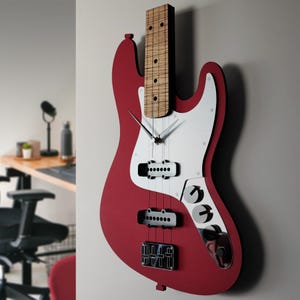 May include: A red bass guitar-shaped wall clock with a white pickguard and silver-colored knobs. The clock has a wooden neck and fretboard, with black pickups and a black bridge. The clock hands are silver.