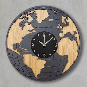 Könnte beinhalten: Eine runde Wanduhr in Form eines Globus. Das Zifferblatt ist schwarz mit weißen Zeigern und goldenen Ziffern. Die Kontinente sind aus grauem Material ausgeschnitten, wodurch ein heller Holzmaserungshintergrund sichtbar wird. Die Uhr ist an einer grauen Wand befestigt.