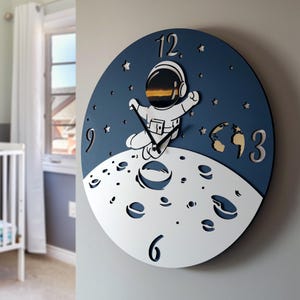 May include: A round wall clock with a navy blue background featuring a white astronaut floating above a white moon. Silver numerals mark the hours. The clock is approximately 12 inches in diameter.