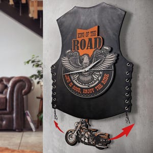 May include: A black wall clock shaped like a leather vest, featuring a motorcycle theme. The clock has an eagle emblem, the words "KING OF THE ROAD" and "LIFE IS GOOD, ENJOY THE RIDE". A motorcycle pendulum hangs below, with red arrow accents.