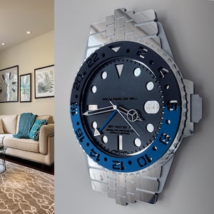 Puede incluir: Un gran reloj de pared con forma de reloj de pulsera. La esfera es azul oscuro con marcadores y agujas blancas. El bisel es azul y plateado, y la correa es plateada. El reloj indica "GMT-MASTER" y "OYSTER PERPETUAL DATE".