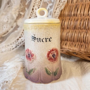 May include: Vintage ceramic sugar jar with lid. The jar is off-white with a cracked glaze, featuring the word "Sucre" in black script. It has red and pink floral designs and a scalloped purple base.