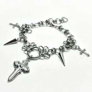 May include: Silver bracelet with a chain link design and various charms. The charms include crosses and cone-shaped spikes. The bracelet has a decorative cross pendant and is set against a white background.