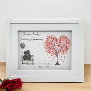 May include: A framed pebble art piece with a heart-shaped tree with red hearts, a couple sitting on a bench, and the text "On your Ruby Wedding Anniversary 40" and "Susan and Andy 29th June 2025".