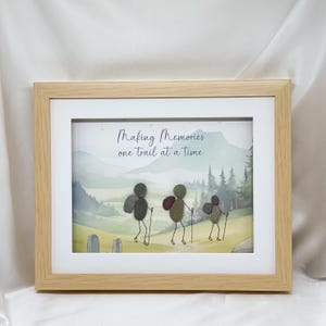 May include: A framed picture with a watercolour landscape scene and three stone figures hiking with walking sticks. The text "Making Memories one trail at a time" is above the figures. The frame is light brown.