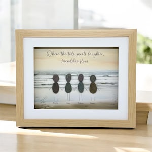May include: Framed artwork depicting four pebble figures holding hands on a beach scene. The text reads "Where the tide meets laughter, friendship flows." Names are written below the figures. The frame is light wood.