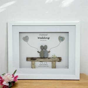 May include: A white framed picture with a pebble art design of a couple sitting on a driftwood bench. The couple are holding silver glitter hearts. The text reads "Diamond Wedding Anniversary" and "Gordon and Muriel Married 23rd March 1963".