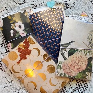 May include: A selection of patterned journals, featuring floral, geometric, and celestial designs. Each journal is held together with an elastic band. A heart-shaped paperclip is visible. Measurements are not available.