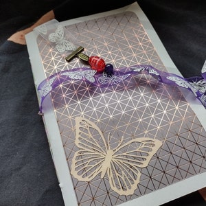 May include: A purple and white ribbon with butterfly designs is tied around a journal with a geometric pattern. A wooden butterfly cutout is attached to the front cover.
