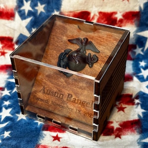 May include: A wooden box with a clear acrylic lid, featuring a black and silver US Marine Corps emblem. The box is engraved with the name "Austin Rangel" and the date "Enlisted November 19, 2022". The box is sitting on a red, white, and blue American flag.