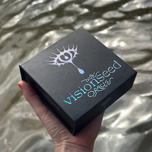 May include: A black square box with the text "the visionseed oracle" in iridescent blue-green lettering. The box features a white and blue eye-shaped design. The box is held in a hand against a blurred water background.